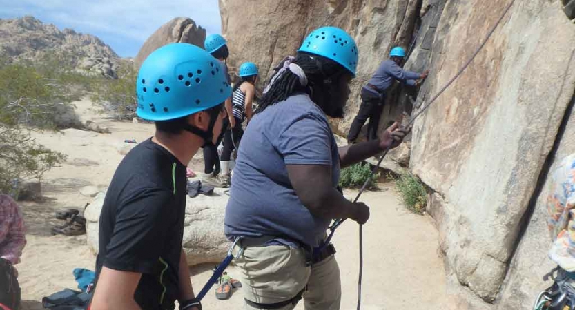 Two people wearing safety gear stand at the bottom of a rock face belaying unseen climbers. 